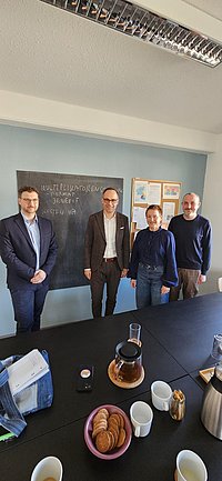 Gruppenfoto von vier Erwachsenen vor einer beschrifteten Tafel bei einer Veranstaltung